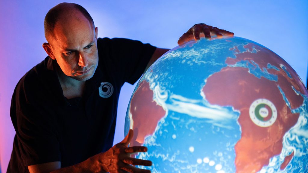 Person with short hair and a dark polo shirt looks closely at a large illuminated globe, in a dark room. They touch the globe with both hands.