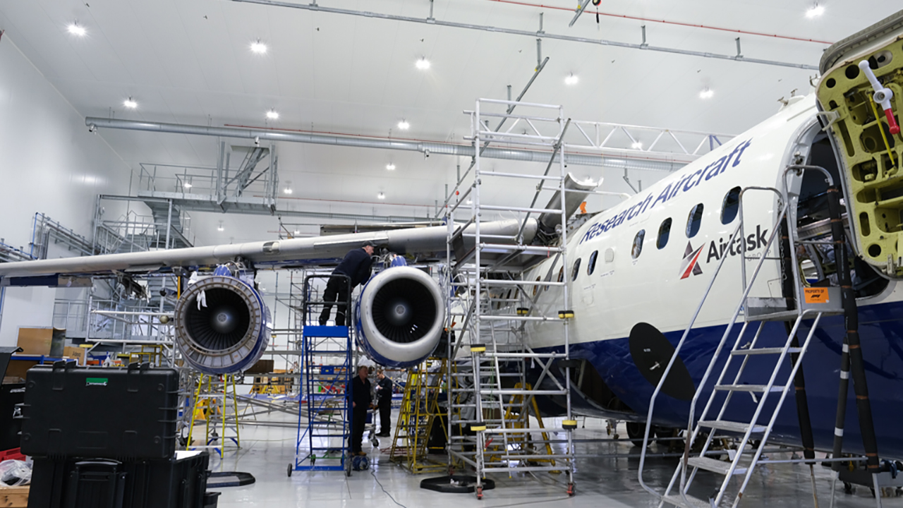 A large blue and white research aircraft in a hangar. It has scaffolding around it and is undergoing maintenance.