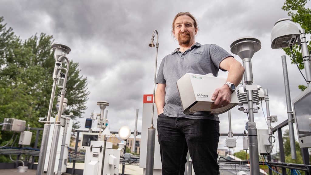 Person with long hair and beard stands on a rooftop observatory holding a white, rectangular scientific instrument under one arm.