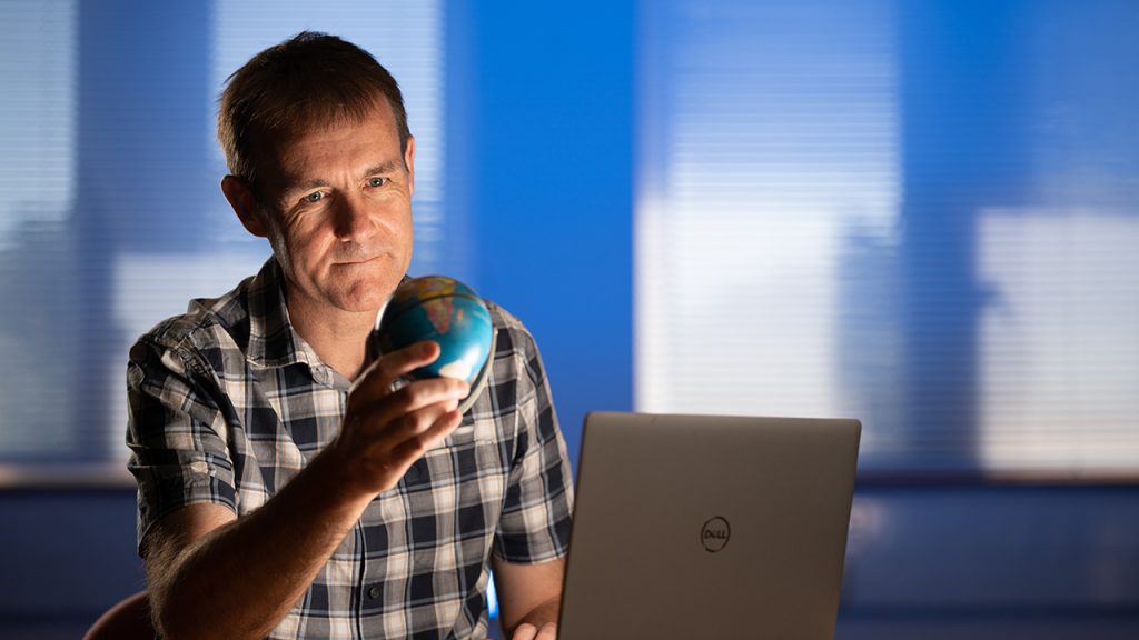 Person wearing a check shirt sits at a desk inside with a laptop on the table. They hold a small globe in one hand, and inspect it.