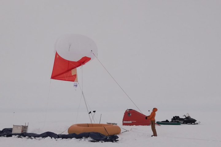 Person standing under orange and white Helikite. Snow and white sky in background.