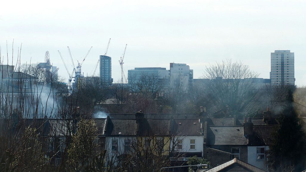 Row of houses and trees In foreground. In the background there are blocks of flats. The sky is grey and there's smoke from chimneys.