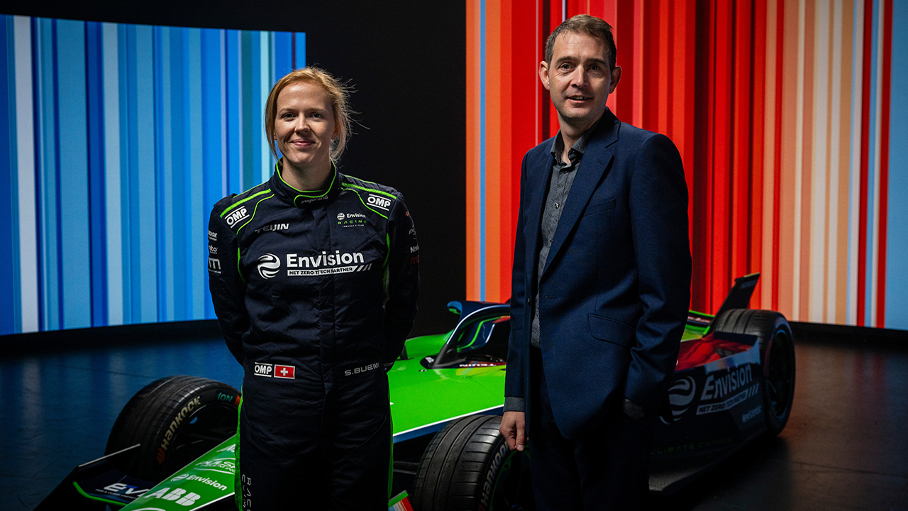 Two people stood in front of a Formula E racing car. Blue and red stripes design on the wall in background.