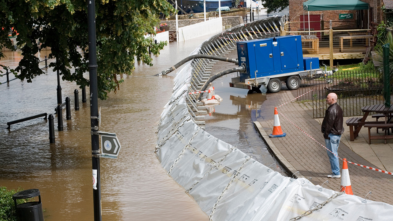 Brown river water floods a pavement and is rising up a grey plastic and metal chained flood defence barrier. A blue machine on wheels with tubes going into the water pumps the flood water away from a building. A person with a bald head wearing a dark leather jacket and blue jeans stands nearby and looks at the brown flood water.