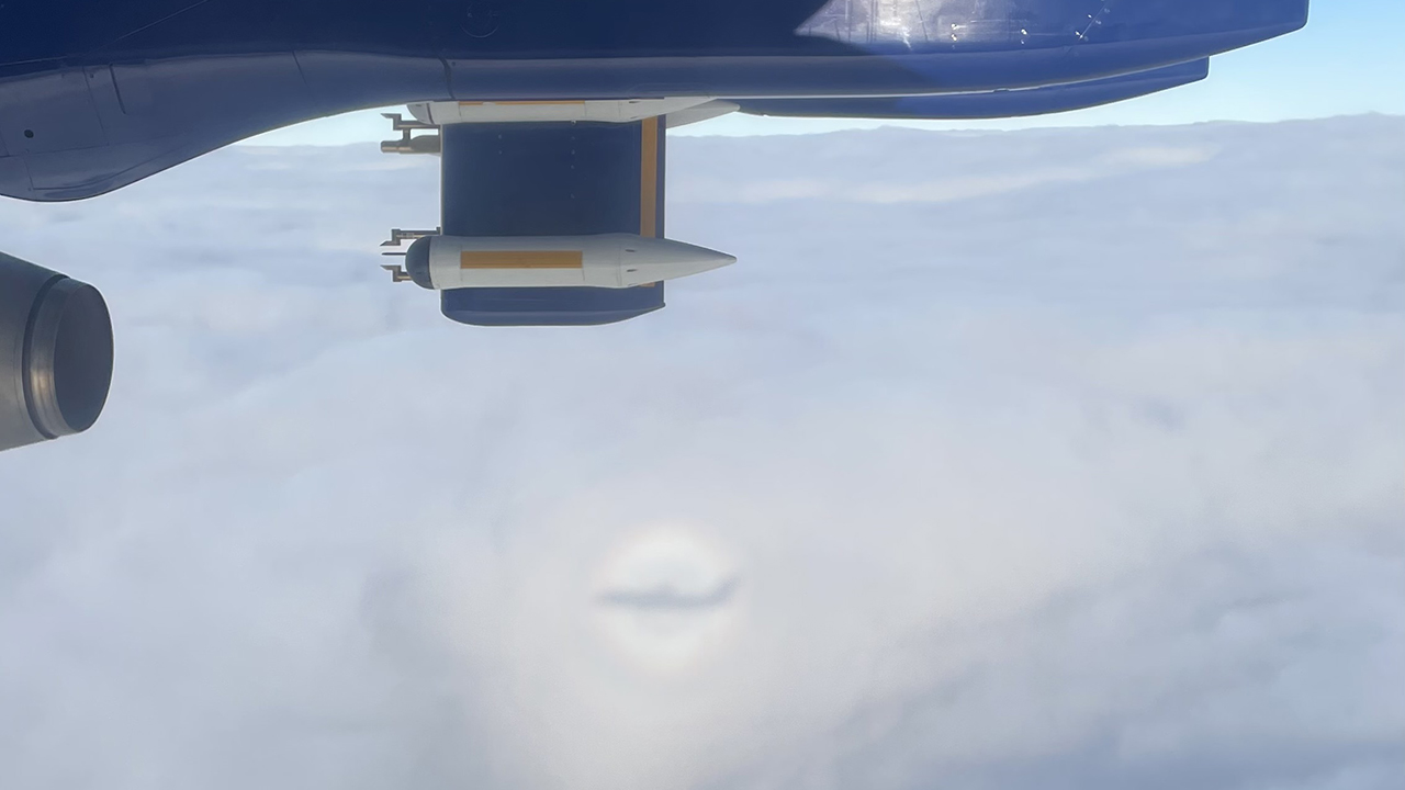 Underside of a blue aircraft wing, above clouds, with a small shadow of the aircraft cast onto the clouds below, with a pastel coloured circular rainbow around the shadow - known as a halo.