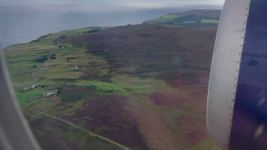View from an aircraft window, of green and purple ground near the sea. A blue and grey aircraft engine is just visible to the right have side of the image.