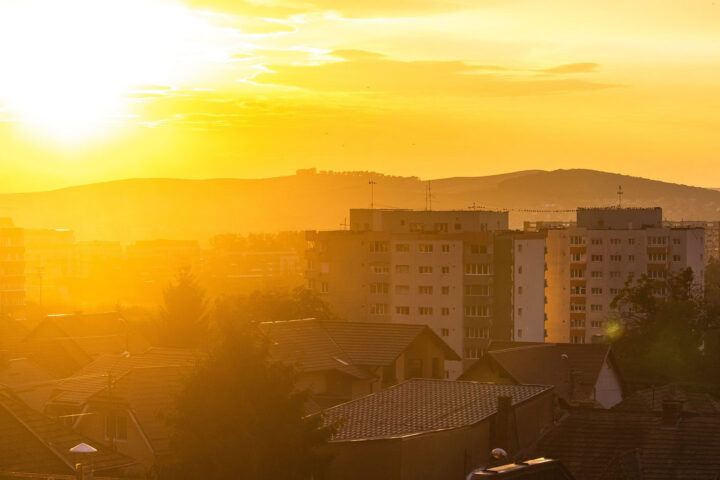 Yellow and orange city skyline with the sun bright in the sky. In the foreground there are houses, trees and flat block towers.