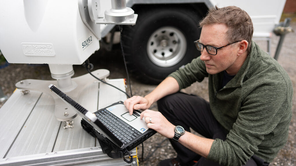Person wearing glasses and green fleece sits on tarmac, operating a laptop connected to a large, white scientific instrument.