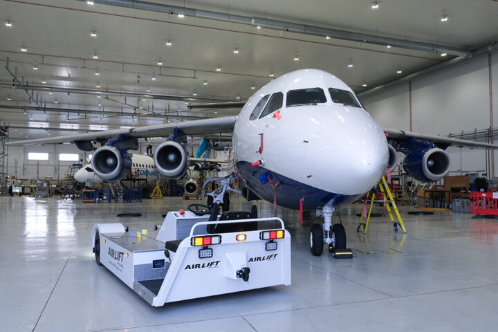An electric tug vehicle sits in front of the nose of a large white and blue aircraft.