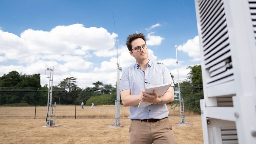 Person wearing glasses and short sleeved shirt stands with a notepad in an outdoors space. They look towards a white scientific instrument box in front of them.