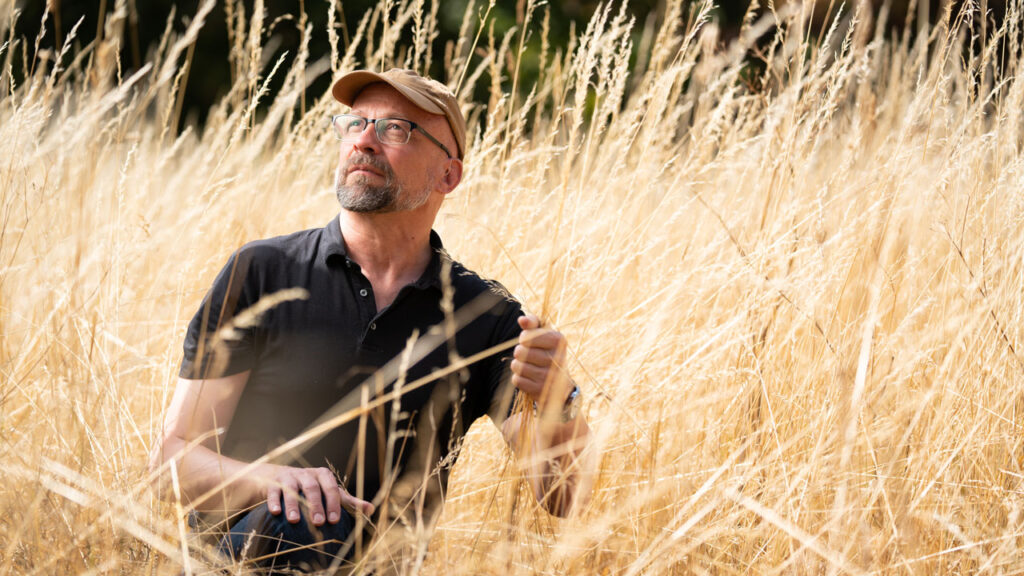 Person with a beard, wearing glasses and a peaked cap crouches in a field of long, dry grass. They look upwards towards the sky, and hold strands of grass in one hand.