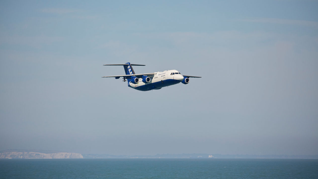 Blue and white aircraft flies low above blue sea. White cliffs in the background.