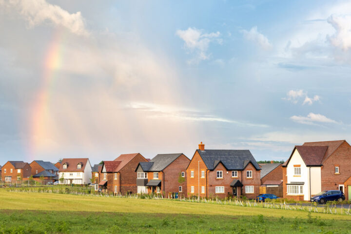 UK housing estate with green grass in foreground and cloudy, blue sky with rainbow in background.