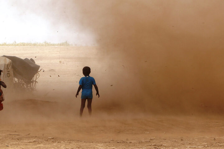 Two children stand in a dusty field. A large dust cloud is blowing around the children.