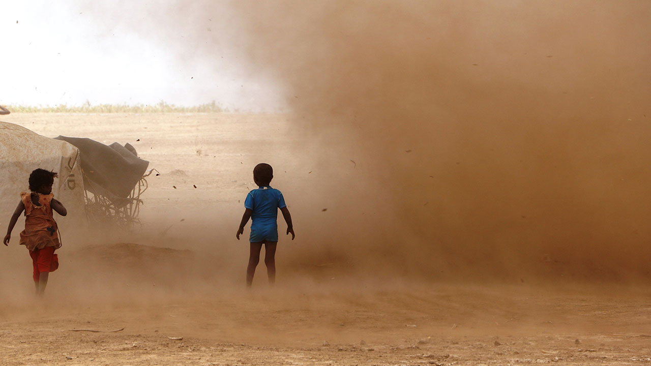 Two children stand in a dusty field. A large dust cloud is blowing around the children.
