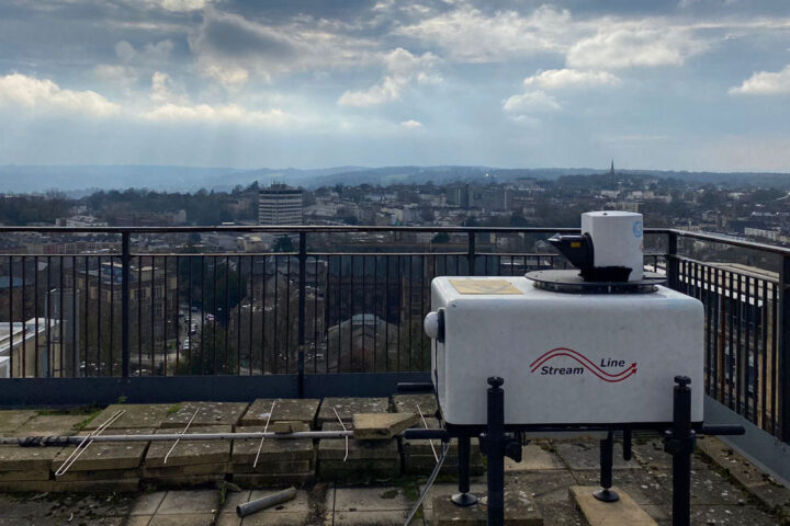 White, square-shaped Lidar instrument on rooftop. Bristol city in background with blue sky and clouds.