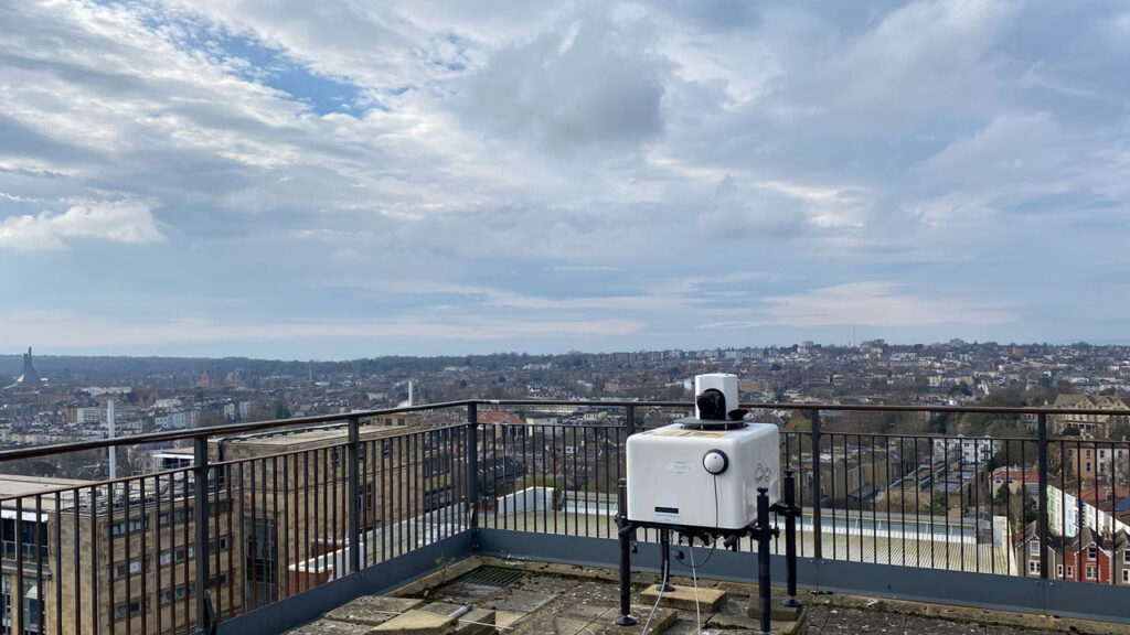 White, square-shaped Lidar instrument on rooftop. Bristol city in background with blue sky and clouds.