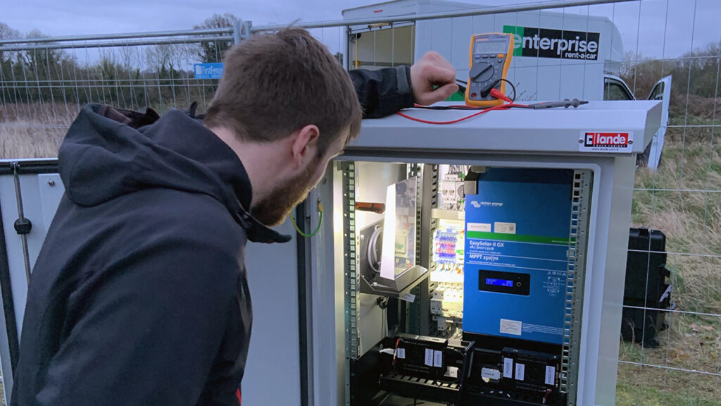 A person with short dark hair wearing a hooded jacket looks inside a white metal box that has electrical components in