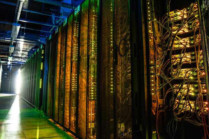 Dark aisle full of racks of computer servers illuminated by small green and blue equipment lights.