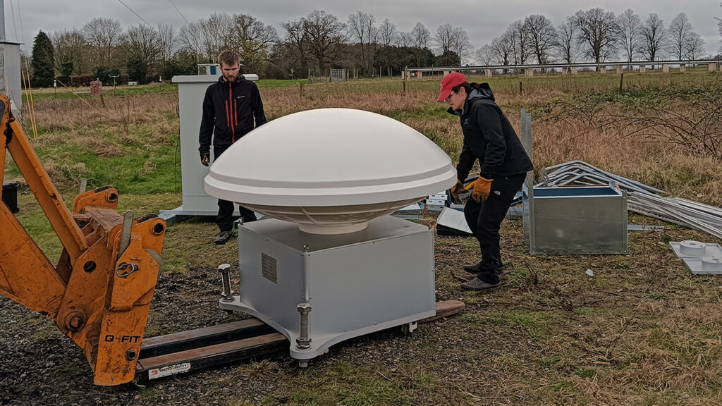 2 people stand either side of a scientific instrument that looks like a white dome on top of a white box.