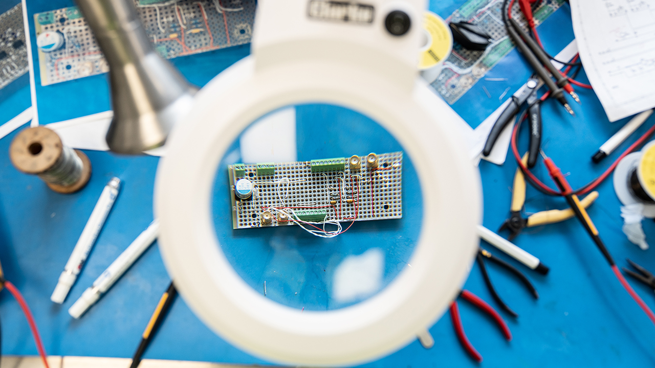 View through a magnifying glass looking at an electrical circuit board on a bench. There are tools scattered around the bench.