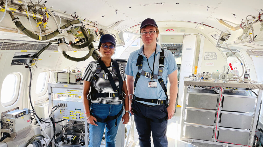 Two people wearing casual clothes and harnesses stand inside an aircraft, which is fitted out with scientific equipment.