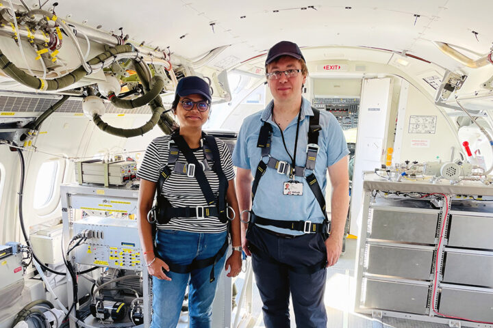 Two people wearing casual clothes and harnesses stand inside an aircraft, which is fitted out with scientific equipment.