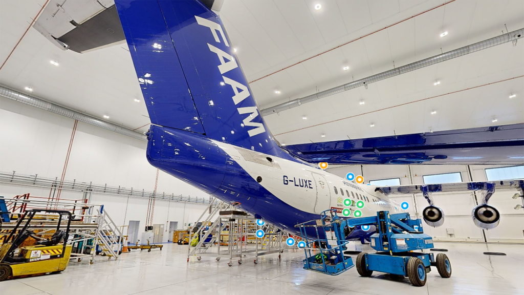 A blue and white aircraft viewed from the tail end, inside a white hangar.