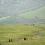 Group of people walking across green grass. Green valley with green trees and houses in the background.