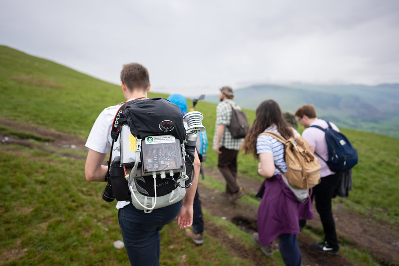 Person carrying a homemade weather monitoring instrument in a backpack. Group of people and green hills in background.