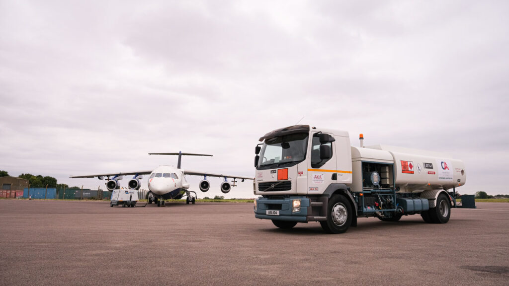 White and blue research aircraft parked outside. Large refuelling truck parked next to the aircraft. Grey sky and clouds in background.