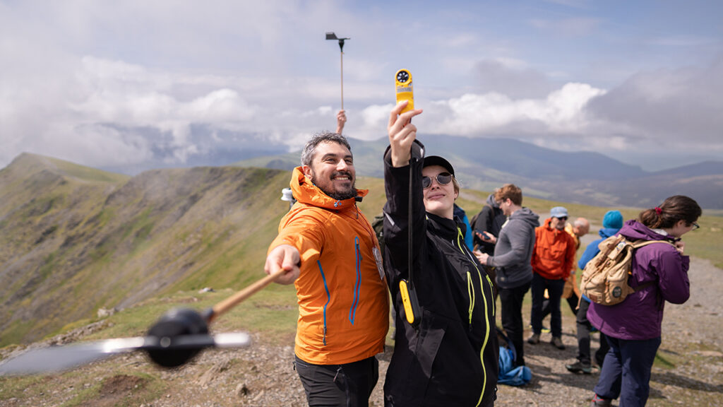 Person wearing orange waterproof holds weather vane. Person wearing black waterproof holds yellow weather monitoring instrument in the air. Group of people stood on gravel path in background with green hills, clouds and blue sky.