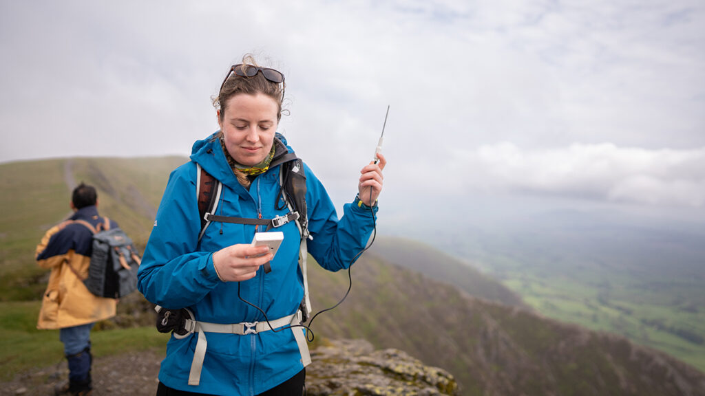 Person wearing blue waterproof coat holds a thermometer in the air. Person stood in the background on rocky grass, with hills and low grey clouds.