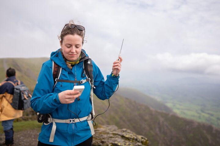 Person wearing blue waterproof coat holds a thermometer in the air. Person stood in the background on rocky grass, with hills and low grey clouds.