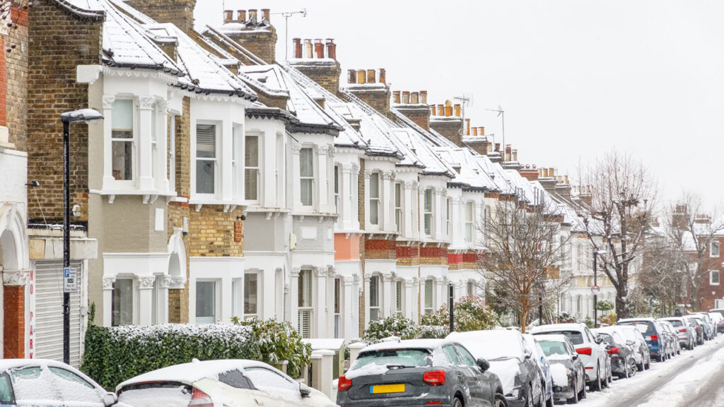 Residential street in the UK in wintertime. Cars are parked in front of houses. The houses, cars and street are covered in snow. Bare trees and grey sky in background.