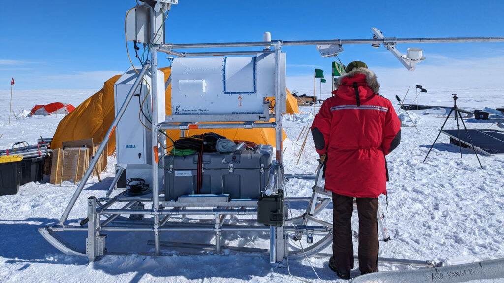 Person wearing red snow coat stood in front of a large scientific instrument on the snow. Blue sky and clouds in background.