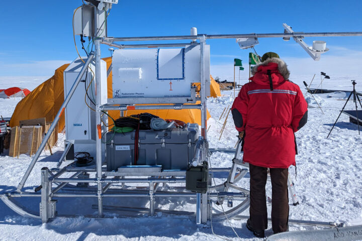 Person wearing red snow coat stood in front of a large scientific instrument on the snow. Blue sky and clouds in background.