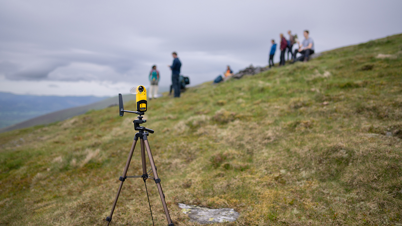 Yellow weather monitoring instrument attached to a tripod. A group of people, green grass and grey sky in background.