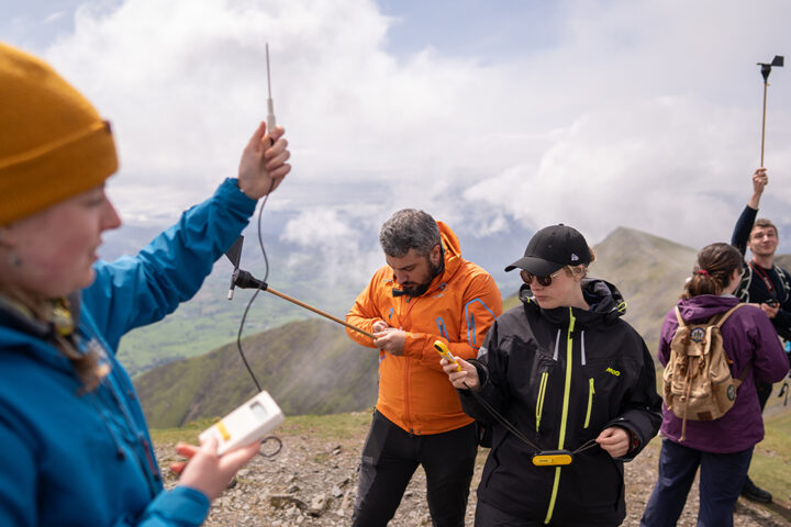 Person in blue raincoat holds up a thermometer. Behind them a group of people are also holding scientific instruments. Green rocky hills, clouds and blue sky in background.