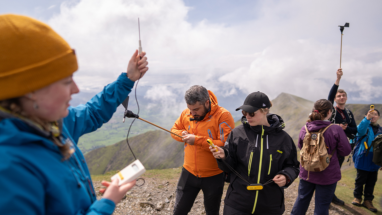 Person in blue raincoat holds up a thermometer. Behind them a group of people are also holding scientific instruments. Green rocky hills, clouds and blue sky in background.