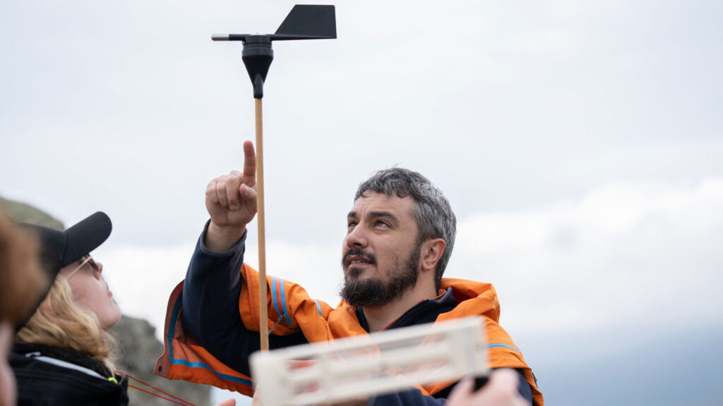 Person wearing orange raincoat is holding a weather vane in the air. They are pointing at the instrument while another person wearing a black raincoat looks at it.