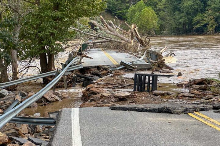 Brown flood waters surround road severely damaged by Hurricane Helene. Fallen trees and debri lie across the road and in the water,