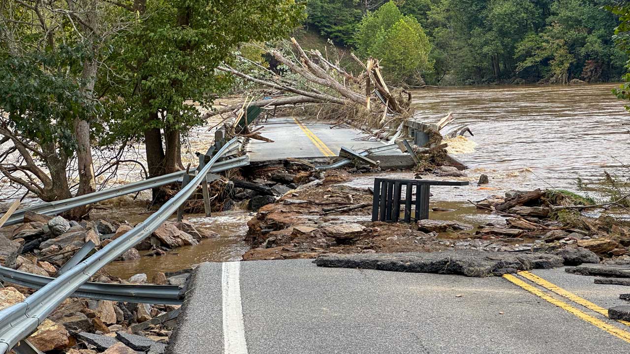 Brown flood waters surround road severely damaged by Hurricane Helene. Fallen trees and debri lie across the road and in the water.