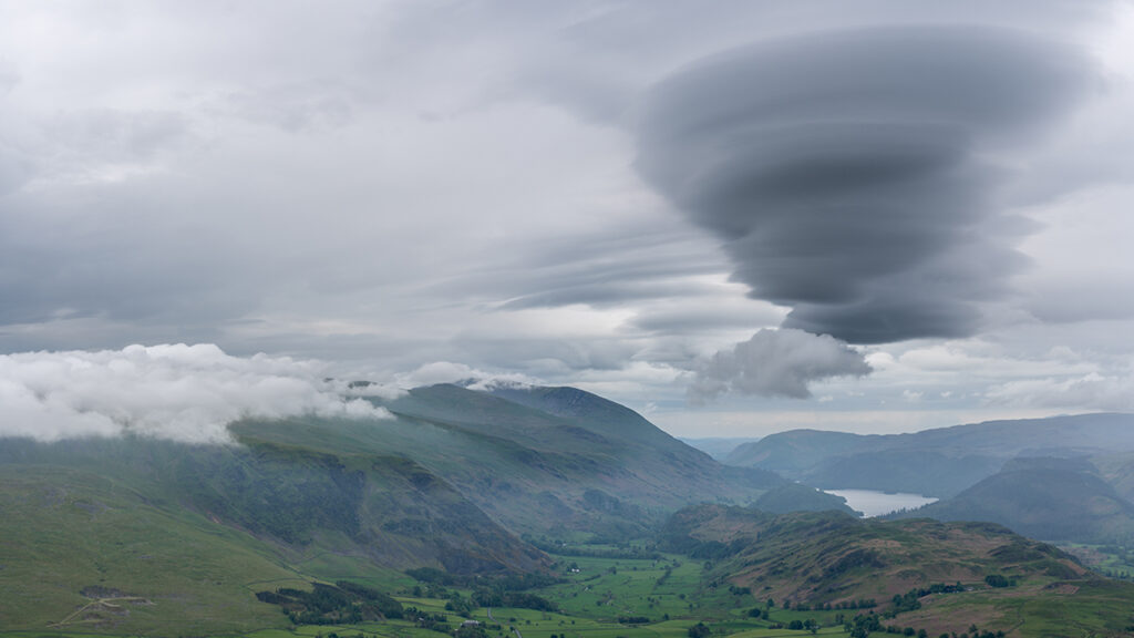 Green mountains and green valley with trees, houses and a lake. Low hanging grey lenticular clouds in the sky.