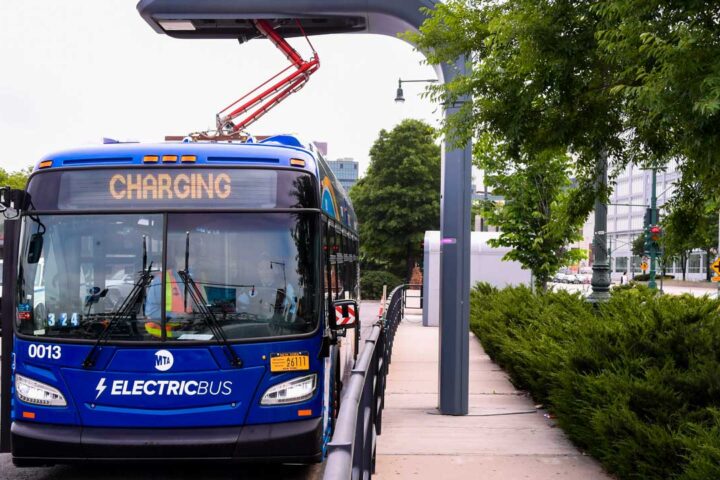 Dark blue electric bus plugged into overhead charging port. Railing and pavement next to bus. Cars, building, hedges and trees in background. Grey sky overhead.