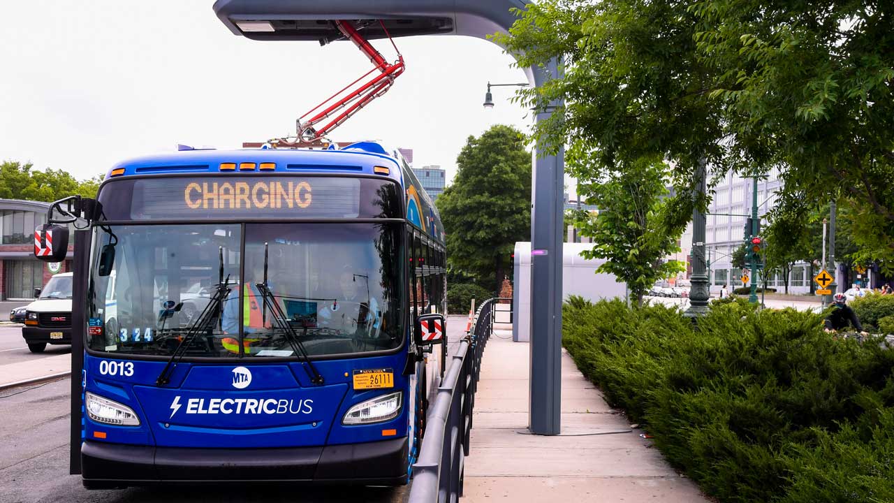 Dark blue electric bus plugged into overhead charging port. Railing and pavement next to bus. Cars, building, hedges and trees in background. Grey sky overhead.