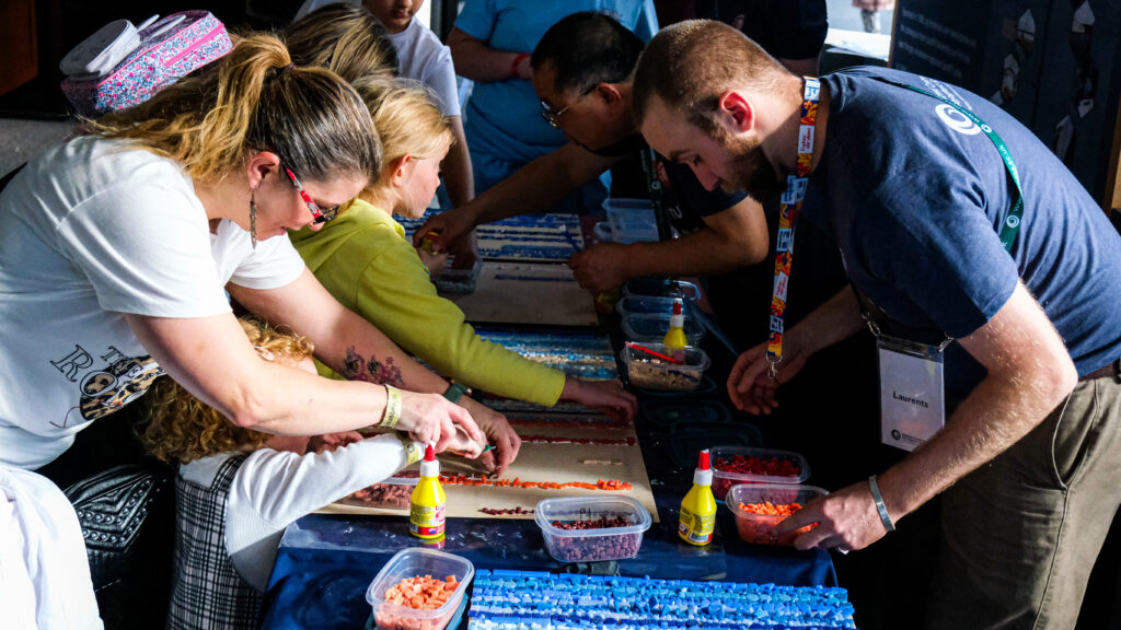 A group of people lean over a long table. They place mosaic tiles in shades of blue and red on a board.