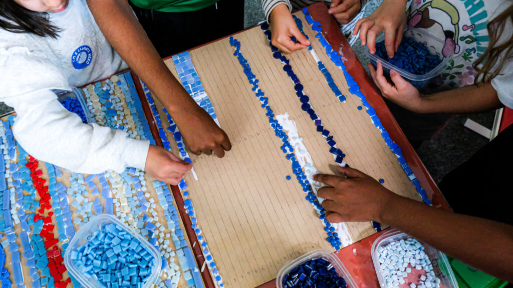 Multiple hands place blue mosaic tiles on a lined board.