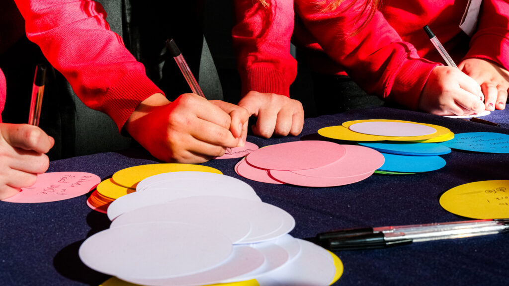 Three sets of hands write on colourful pieces of paper. There are pens and more paper pieces scattered in front of them.