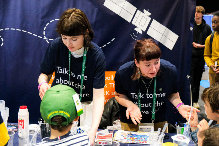 Two people wearing t-shirts that say "Talk to me about the air we share", talking to a group of children. There is a banner featuring a satellite illustration behind them.